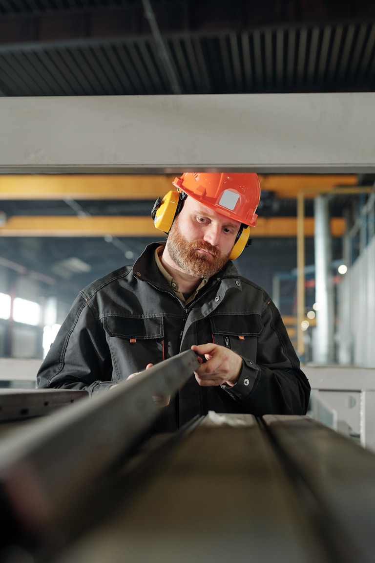 A construction worker in an orange hard hat and yellow earmuffs inspecting a metal beam in an industrial setting.