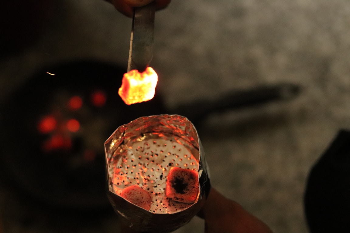 A pair of metal tongs holding a single glowing red hookah charcoal cube above a foil-covered bowl.